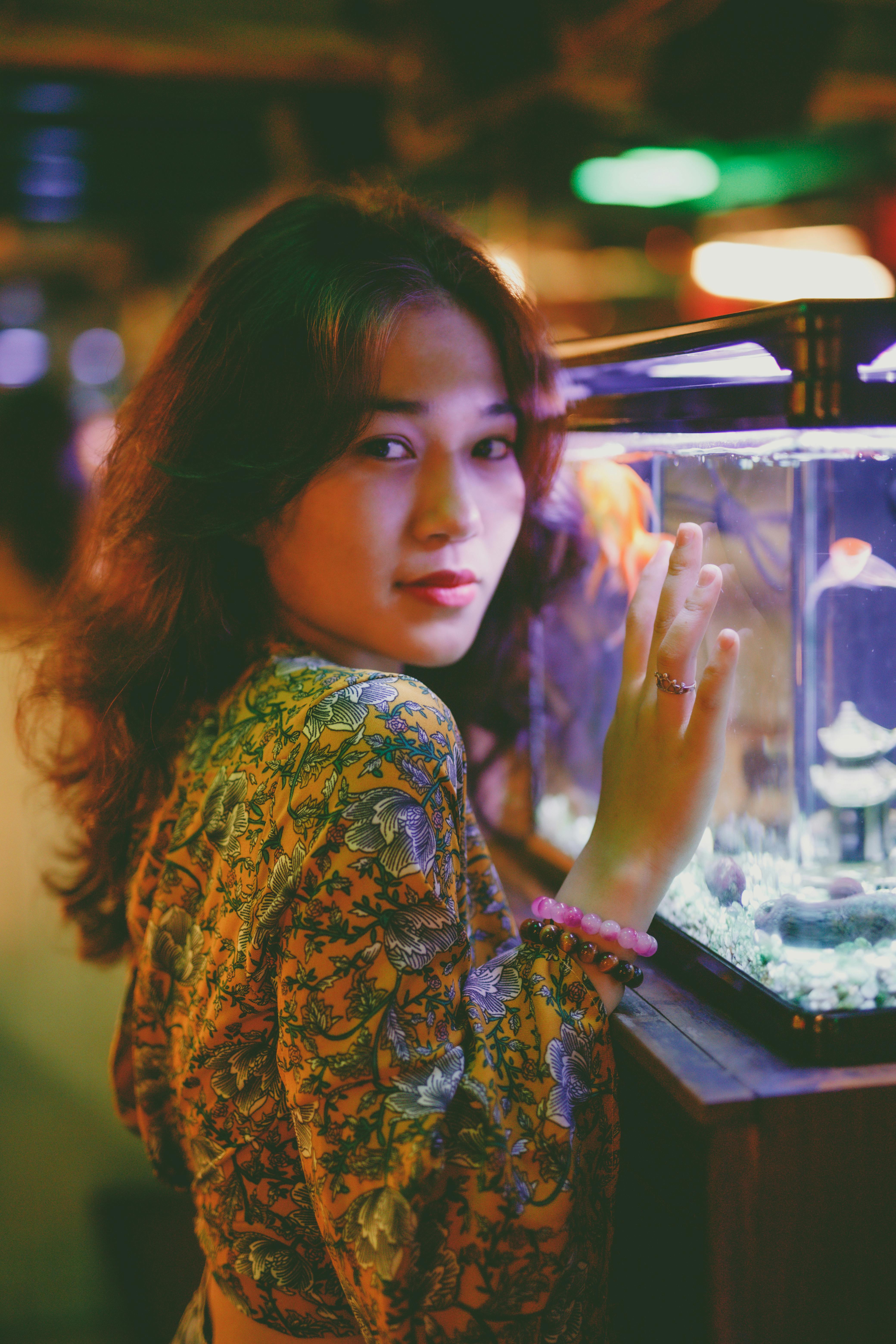 Asian woman in floral top poses by a lit aquarium, capturing serene beauty.