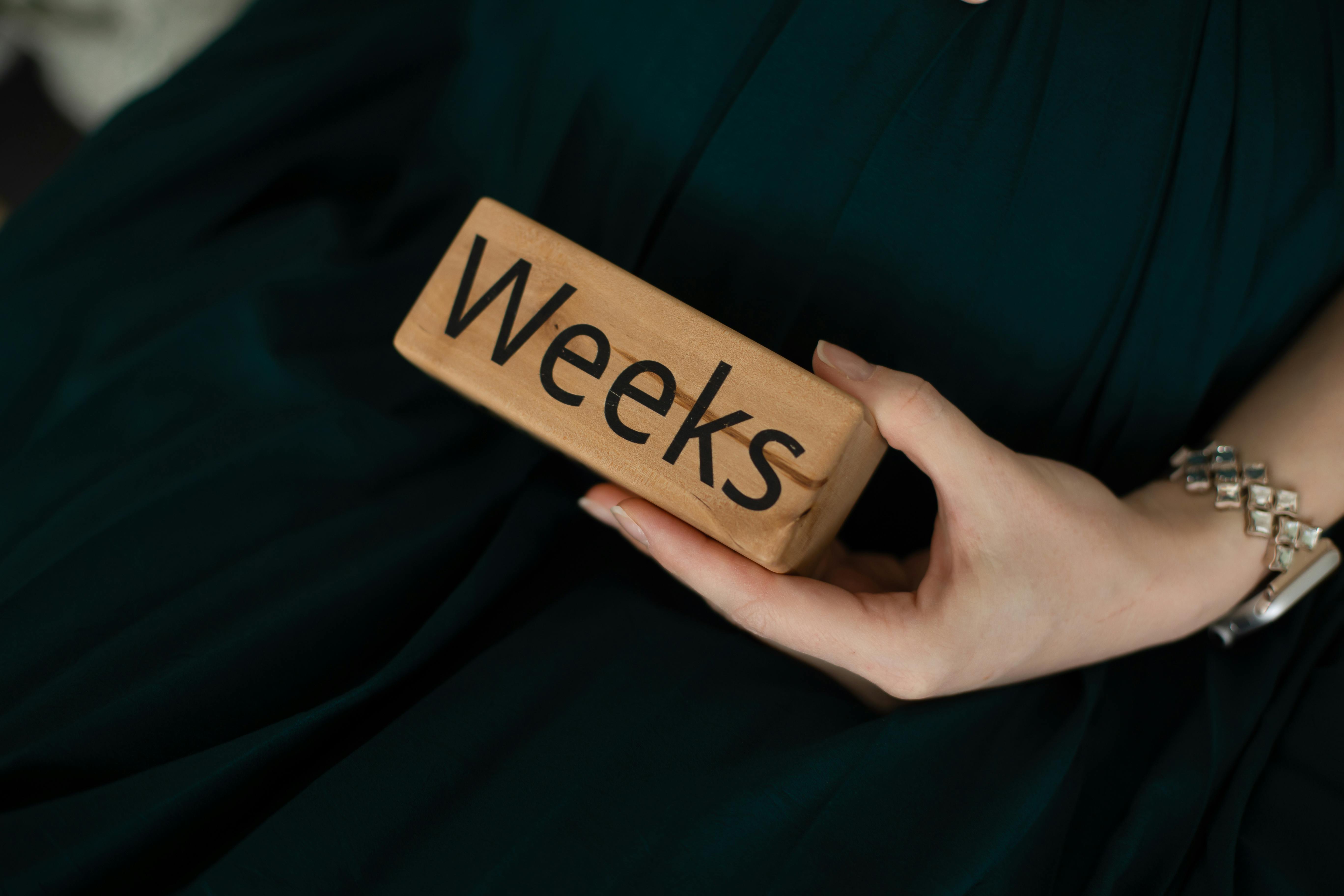 Close-up of a woman's hand holding a wooden block with the text 'Weeks,' on a green background.