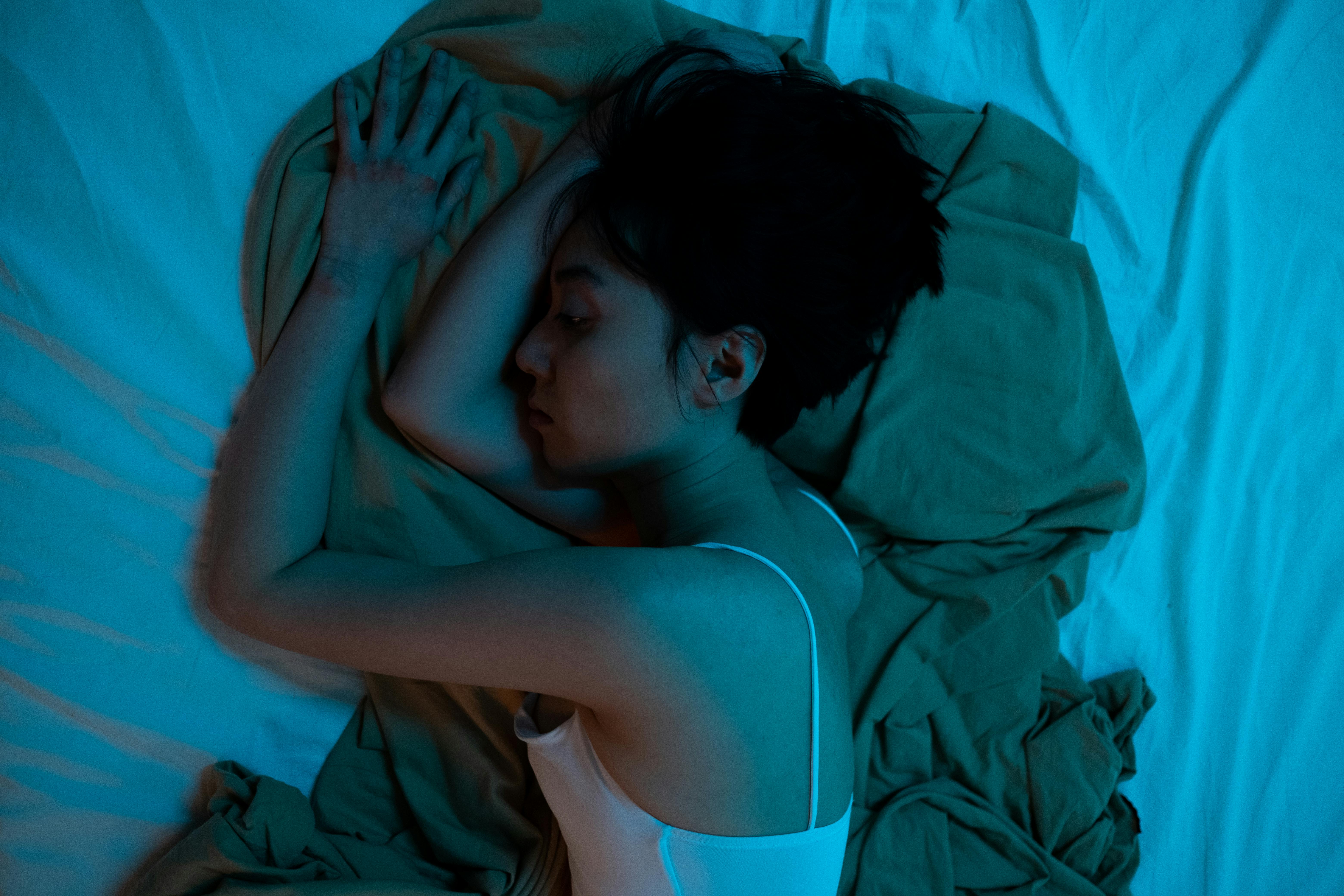 Asian woman lying on bed, expressing a contemplative mood, blue lighting.