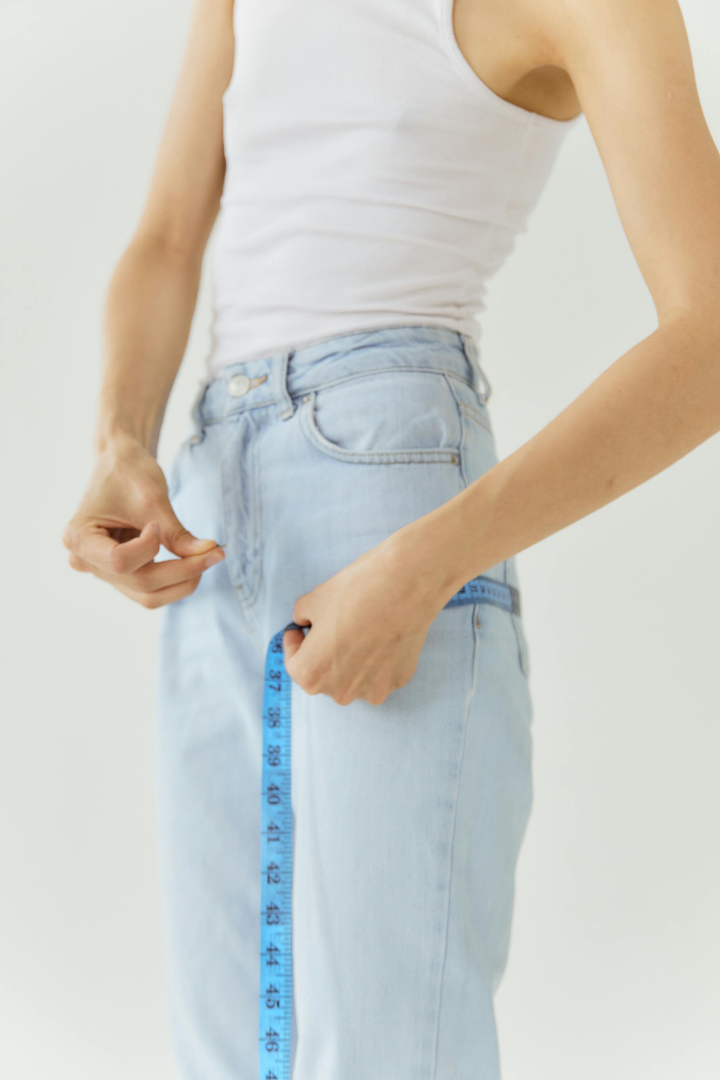 Person wearing denim jeans and white tank top uses a tape measure for size checking.